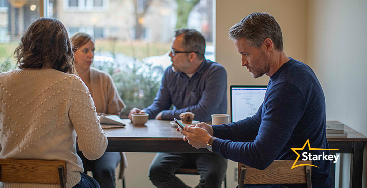 Man sitting at coffee shop table with 3 friends looking at smartphone in hand
