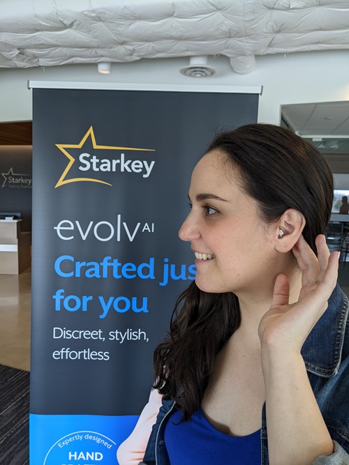 April Corner standing in front of Evolv AI pull-up banner holding left hand up to left ear